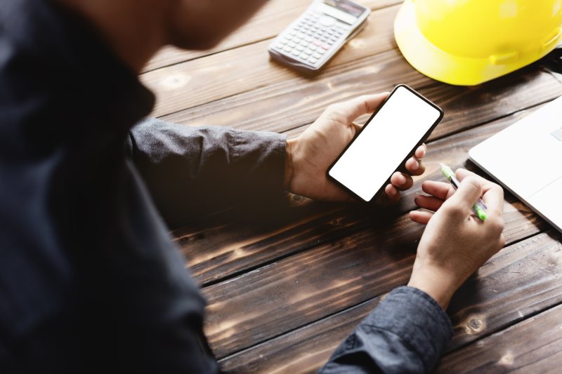 Worker holding smartphone with blank screen at wooden desk with calculator and hard hat